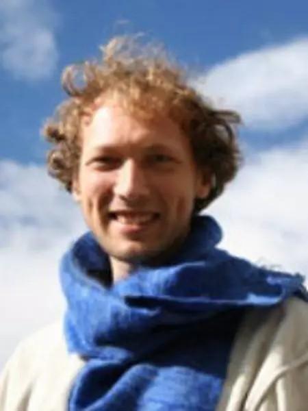 A person with curly hair is smiling outdoors, wearing a blue scarf and light-colored clothing, with a partly cloudy sky in the background.