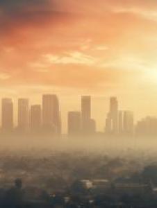 Hazy city skyline at sunrise with tall buildings partly obscured by smog and dense clouds, casting a warm glow over the urban landscape.