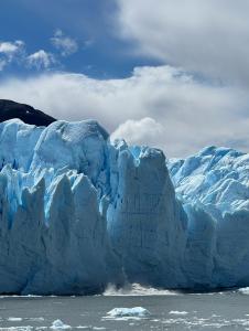 Large blue glacier with jagged ice peaks and a chunk breaking off into the water below, under a partly cloudy sky.