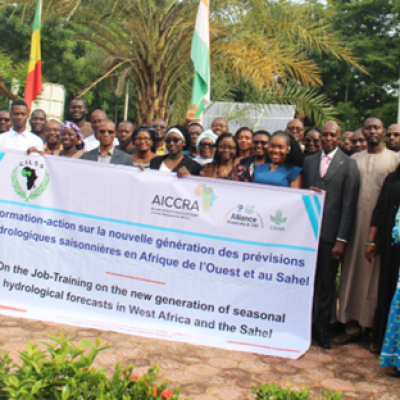 A large group of people pose outdoors, holding a banner about hydrological forecast training in West Africa and the Sahel, with flags and trees in the background.