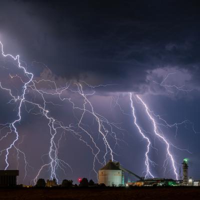 Multiple lightning bolts strike the ground near industrial buildings under a dark, cloudy night sky.
