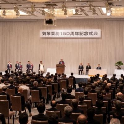 A formal ceremony in a large hall with seated attendees, a speaker at the podium, and banners in Japanese, alongside the Japanese flag and another flag.