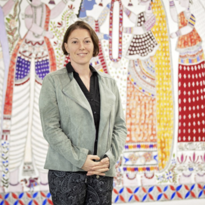 A woman stands indoors in front of a colorful, patterned mural featuring stylized figures in traditional attire.