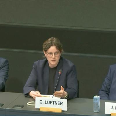 Three people sit at a panel table, with microphones and nameplates in front of them. The person in the center is speaking while the others listen.