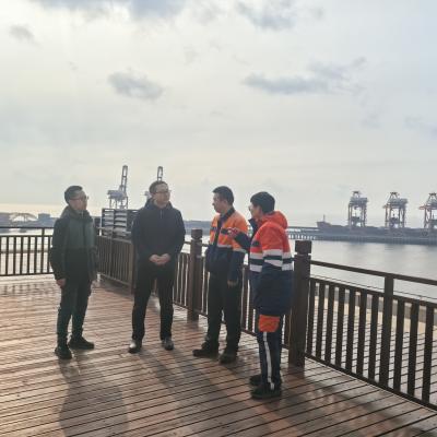 Four people stand on a wooden deck by the water with cranes and industrial structures visible in the background under a cloudy sky.