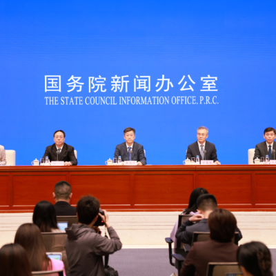 Five officials sit at a long table during a press conference, with "The State Council Information Office, P.R.C." displayed in English and Chinese on a blue screen behind them.