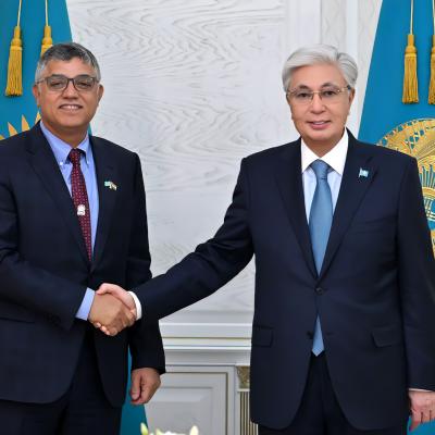 Two men in suits shake hands in front of two blue flags with gold emblems in a formal setting.