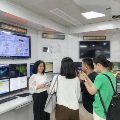 A woman stands in front of several monitors displaying weather data as she is interviewed and photographed by a group of people in a control room.